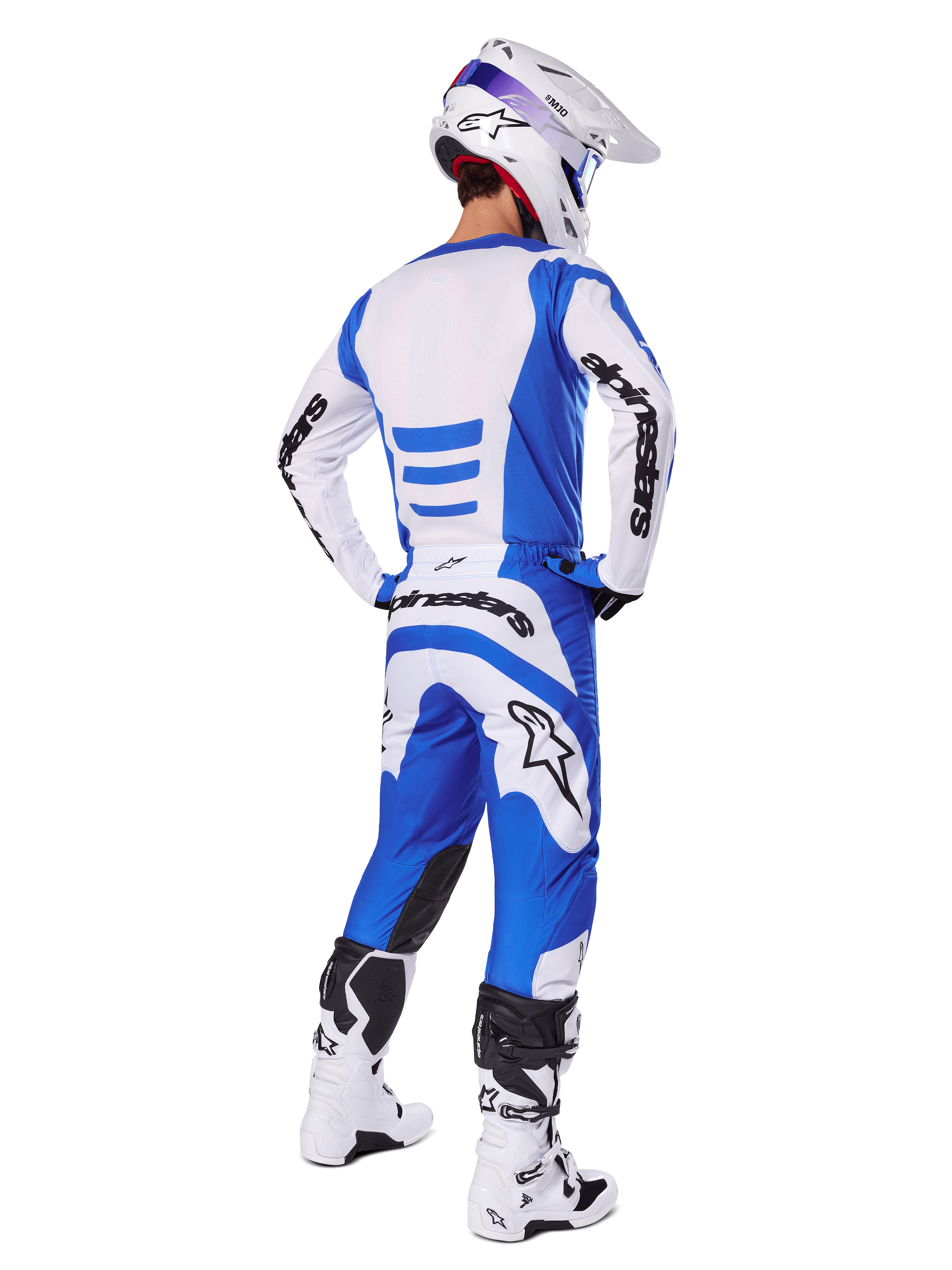 Pilote de motocross de dos portant l'ensemble d'équipement Alpinestars Fluid Haul 2025, maillot bleu et blanc avec pantalon assorti, casque blanc avec accents bleus et bottes de course blanches et noires, tenue de course tout-terrain coordonnée
