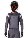 Alpinestars 2025 Fluid Haul Jersey en Gear Set, motorcross outfit, grijs met zwarte en witte accenten, lange mouwen en opvallende branding, zwarte helm met oranje bril en zwarte handschoenen, compleet tenue voor off-road racen
