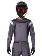 Alpinestars 2025 Fluid Haul Jersey en Gear Set, motorcross outfit, grijs met zwarte en witte accenten, lange mouwen en opvallende branding, zwarte helm met oranje bril en zwarte handschoenen, compleet tenue voor off-road racen