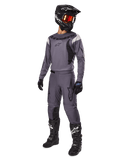 Pilote de motocross portant l'ensemble d'équipement Alpinestars Fluid Haul 2025 en gris, comprenant un maillot et un pantalon gris avec des accents noirs, un casque noir avec des lunettes rougeâtres, des gants noirs et des bottes noires avec des logos blancs, tenue de course tout-terrain complète