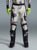 Pantalon Alpinestars Racer Lahnd 2026, pantalon de motocross, motif camouflage sable avec graphismes abstraits gris et violets, porté avec des gants jaune fluo et des bottes noires, avec des logos étoiles noirs sur les cuisses et une ceinture noire à la taille, équipement de course tout-terrain durable