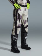 Alpinestars 2026 Racer Lahnd Pants, pantalon de motocross, Sand Camo, porté avec des gants jaune fluo et des bottes de motocross noires, présentant un motif camouflage abstrait avec des panneaux latéraux noirs, design tout-terrain ergonomique