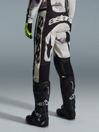 Alpinestars 2026 Racer Lahnd Pants, pantalon de motocross, Sand Camo et gris, vue arrière avec logo blanc imposant sur l'assise et les jambes, associé à des bottes de motocross noires et des gants jaunes, équipement de course tout-terrain professionnel