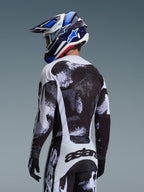 Alpinestars 2026 Racer Lahnd Jersey in Iron Camo, rear view of a motocross rider in a black, white, and grey abstract pattern gear set, including matching pants, black gloves, and a blue racing helmet with goggles.
