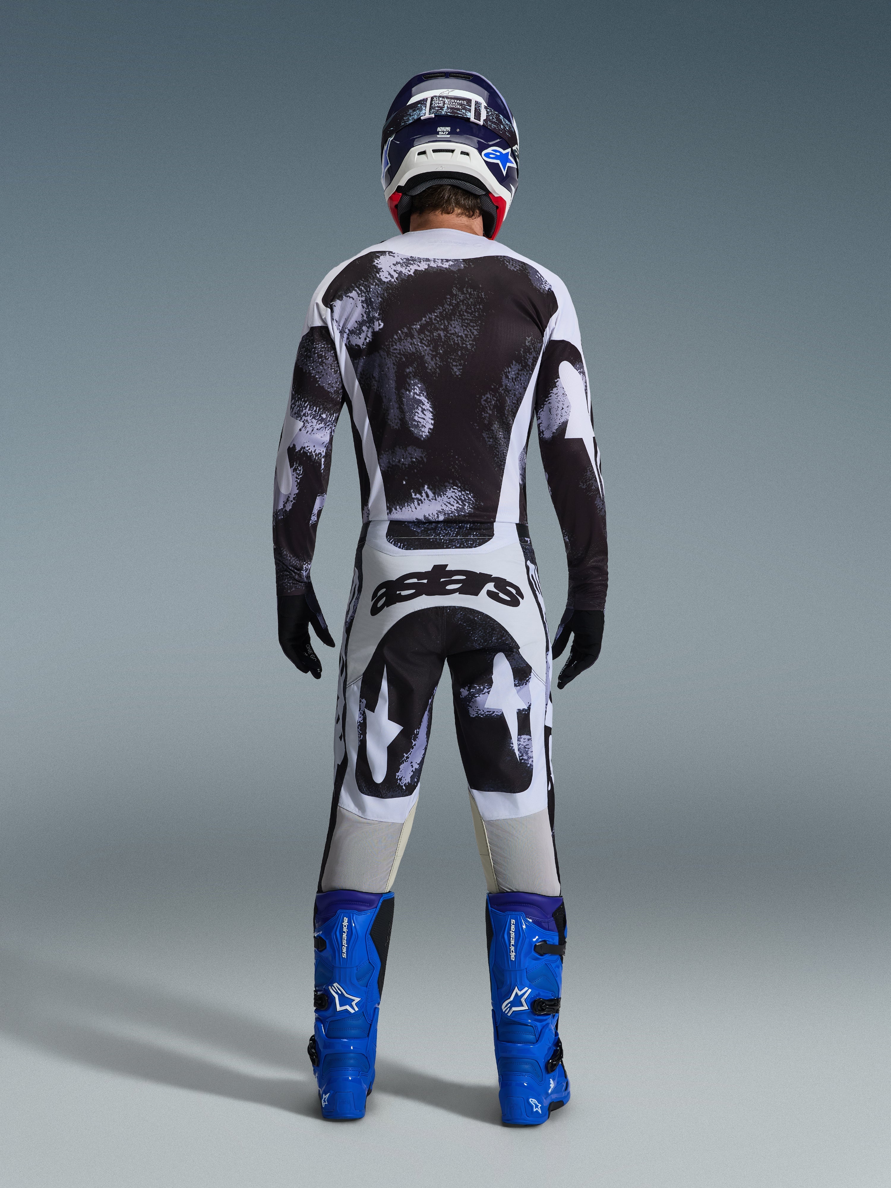 Motocross-Fahrer in der Rückansicht im Alpinestars 2026 Racer Lahnd Gear Set, schwarz-weiß abstrakt gemustertes Jersey und Hose, „astars“-Branding auf dem Gesäß, kombiniert mit blauen Stiefeln und passendem Helm, professionelle Offroad-Rennbekleidung.