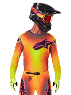 Motocross-Fahrer im 2025 Supertech Lipan Trikot, leuchtender Farbverlauf in Gelb, Orange und Pink, großes schwarzes Alpinestars Stern-Logo mit pinker Kontur auf der Brust, lasergeschnittene Perforationen zur Belüftung, athletische Rennpassform, kombiniert mit passender Hose, gelben Handschuhen und einem schwarzen Helm mit Brille.