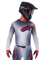 Maillot Alpinestars Supertech Lipan 2025, maillot de motocross, gris avec des accents orange et bleu marine, haut de performance à manches longues avec détails de ventilation perforés, porté avec un casque noir et des gants rouges, design de course épuré
