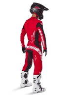 Motocross-Fahrer im Alpinestars 2025 Techstar Dreem Ausrüstungsset in Hellrot und Schwarz, mit weißem Branding auf Trikot und Hose, kombiniert mit einem schwarzen Helm, weiß-schwarzen Stiefeln und schwarz-weißen Handschuhen, Rückansicht in Dreiviertel-Perspektive.