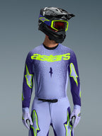 Frontalansicht eines Motocross-Fahrers im 2026 Supertech Scenz Gear Set, hellviolettes und dunkelblaues Trikot mit leuchtend neongelbem 'astars' Branding, passender hellvioletter Hose und Handschuhen mit Neon-Akzenten, schwarzer Helm mit neongelber Brille, Studioaufnahme vor grauem Hintergrund.