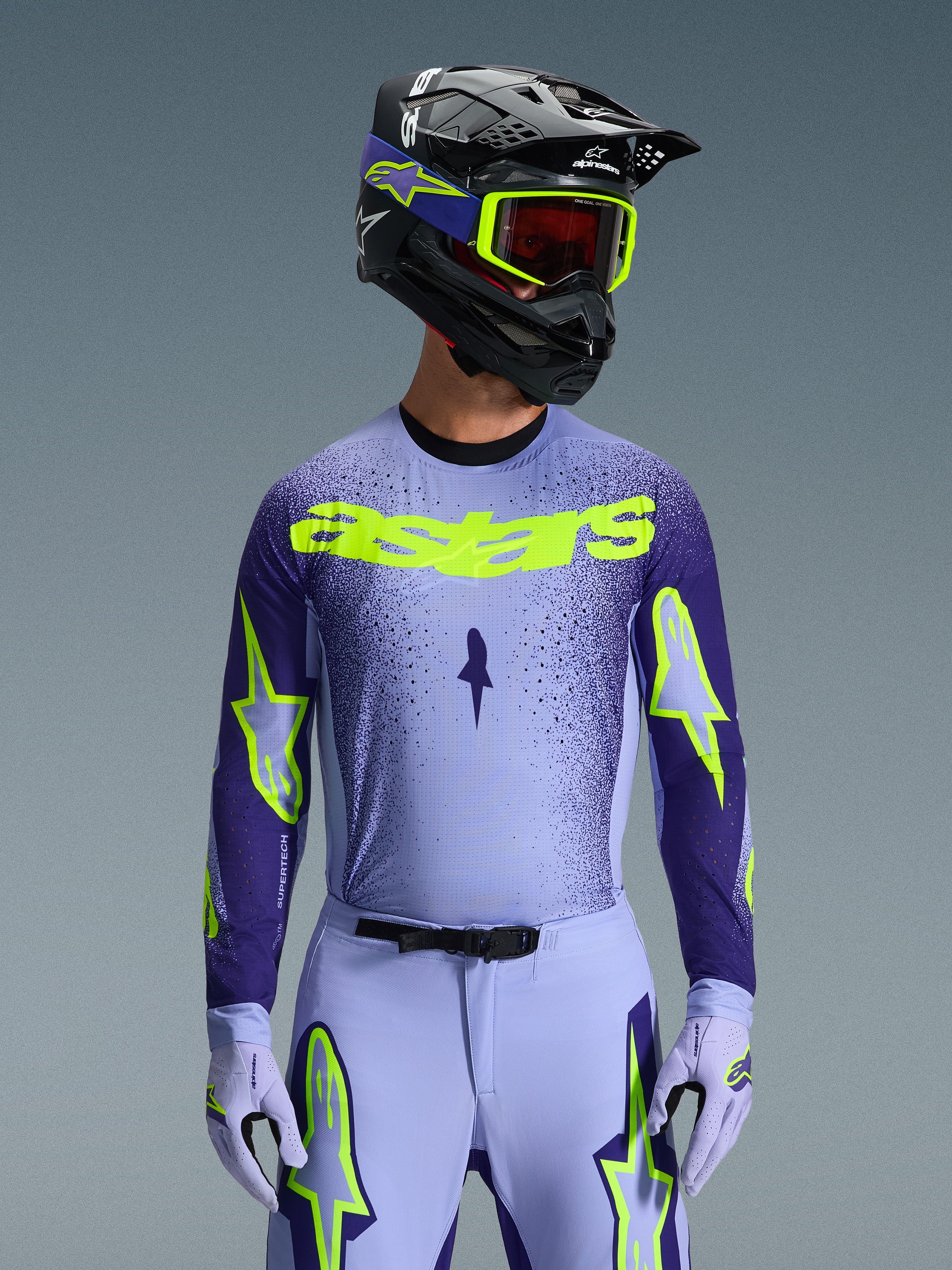 Frontalansicht eines Motocross-Fahrers im 2026 Supertech Scenz Gear Set, hellviolettes und dunkelblaues Trikot mit leuchtend neongelbem 'astars' Branding, passender hellvioletter Hose und Handschuhen mit Neon-Akzenten, schwarzer Helm mit neongelber Brille, Studioaufnahme vor grauem Hintergrund.