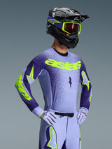 Motorcrosser gekleed in de Alpinestars Supertech Scenz Jersey 2026, paars en neongeel, bijpassende paarse broek met neon accenten, zwarte helm met gele crossbril en paarse racehandschoenen, gecoördineerde professionele uitrusting voor off-road racing