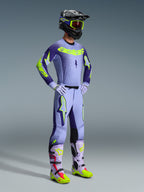 Alpinestars 2026 Supertech Scenz Gear Set, Motocross-Fahrer in kompletter Rennausrüstung, hellviolettes Trikot mit dunkelvioletten Ärmeln und neongelben Akzenten, passende lavendelfarbene Hose, schwarzer Helm mit neongelber Brille und lavendelfarbene Stiefel mit neongrünen und roten Details, professionelles Offroad-Outfit.