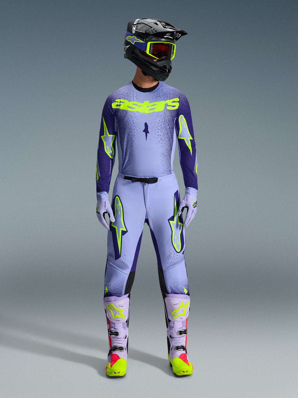 Motocross-Fahrer in kompletter Alpinestars 2026 Supertech Scenz Rennausrüstung, Trikot in Lila und Gelb Fluo mit passender Hose, schwarzer Helm mit gelber Schutzbrille, helllila und neonfarbene Stiefel, professionelles Outfit mit athletischer Passform für den Offroad-Rennsport