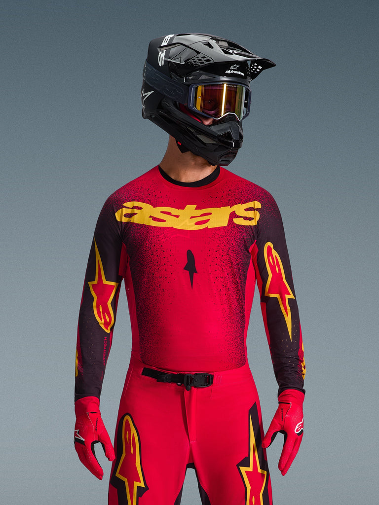 Alpinestars 2026 Supertech Scenz Jersey, motorcross jersey, rood, zwart en geel, gedragen door een rijder met bijpassende zwarte helm en rode handschoenen, voorzien van een opvallend geel 'astars' logo op de borst en een zwart gespikkeld verloop-patroon over de schouders.