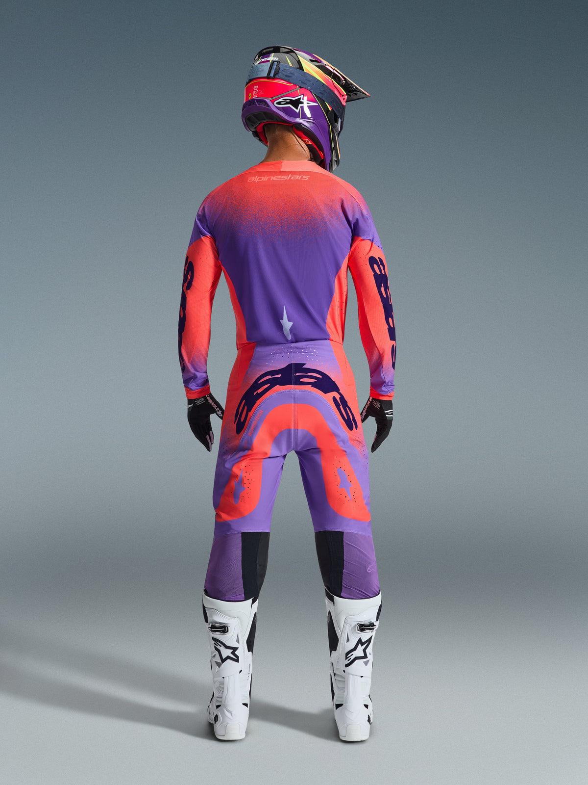Motocross rider from behind wearing the 2026 Supertech Scenz Gear Set in Hot Coral Purple, vibrant jersey and pants with gradient transition, featuring Alpinestars logos, matching helmet, white and black boots, and black gloves, professional gear for elite off-road racing.