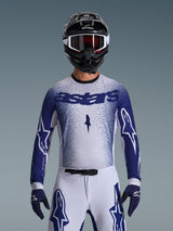Motorcrosser die de Alpinestars 2026 Supertech Scenz Jersey draagt, lichtgrijs en donkerblauw met een gespikkeld gradiëntontwerp en opvallende 'astars' branding. Gedragen met bijpassende witte motorcrossbroek, donkerblauwe handschoenen en een zwarte helm met crossbril, gecoördineerde outfit voor professioneel off-road racen.