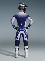 Motorcrossrijder van achteren gezien in de Alpinestars 2026 Supertech Scenz Jersey, lichtgrijs en donkerblauw met een gespikkeld kleurverloop, bijpassende motorcrossbroek, witte racelaarzen en zwarte helm, complete off-road race-outfit.