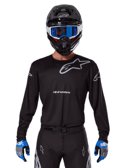 Maillot Alpinestars Racer Graphite 2025, équipement de motocross, noir avec logos gris, porté avec un casque noir assorti, des lunettes bleues et des gants bleus et gris, design de performance ajusté avec marquage sur la poitrine et les bras