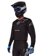 Motocross rider wearing Alpinestars 2025 Racer Graphite Gear Set, Black jersey with grey Alpinestars logos, matching black pants with grey accents, black helmet with blue and white goggles, blue and grey gloves, sleek technical outfit for off-road racing
