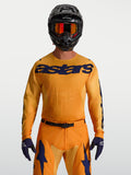 Alpinestars Racer Amber Trikot, Motocross-Renntrikot für Herren, Orange und Dunkelblau, feuchtigkeitsableitendes Material, getragen von einem Model mit Helm und Handschuhen.