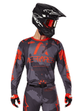 Alpinestars 2025 Racer Hollow Jersey, motorcross jersey, grijs camo en fluo oranje, gedragen met bijpassende kledingset inclusief broek en handschoenen, met een zwarte helm met witte graphics en crossbril, opvallende ASTARS-branding, beschermende off-road race-sportkleding