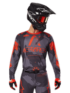 Alpinestars 2025 Racer Hollow Jersey, motorcross jersey, grijs camo en fluo oranje, gedragen met bijpassende kledingset inclusief broek en handschoenen, met een zwarte helm met witte graphics en crossbril, opvallende ASTARS-branding, beschermende off-road race-sportkleding