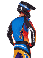 Pilote de motocross vu de dos portant l'ensemble d'équipement Alpinestars 2025 Fluid Wurx, maillot multicolore avec dos bleu et manches noires arborant des logos blancs, pantalon assorti orange et bleu, casque noir et bleu avec lunettes orange, tenue de course tout-terrain coordonnée.