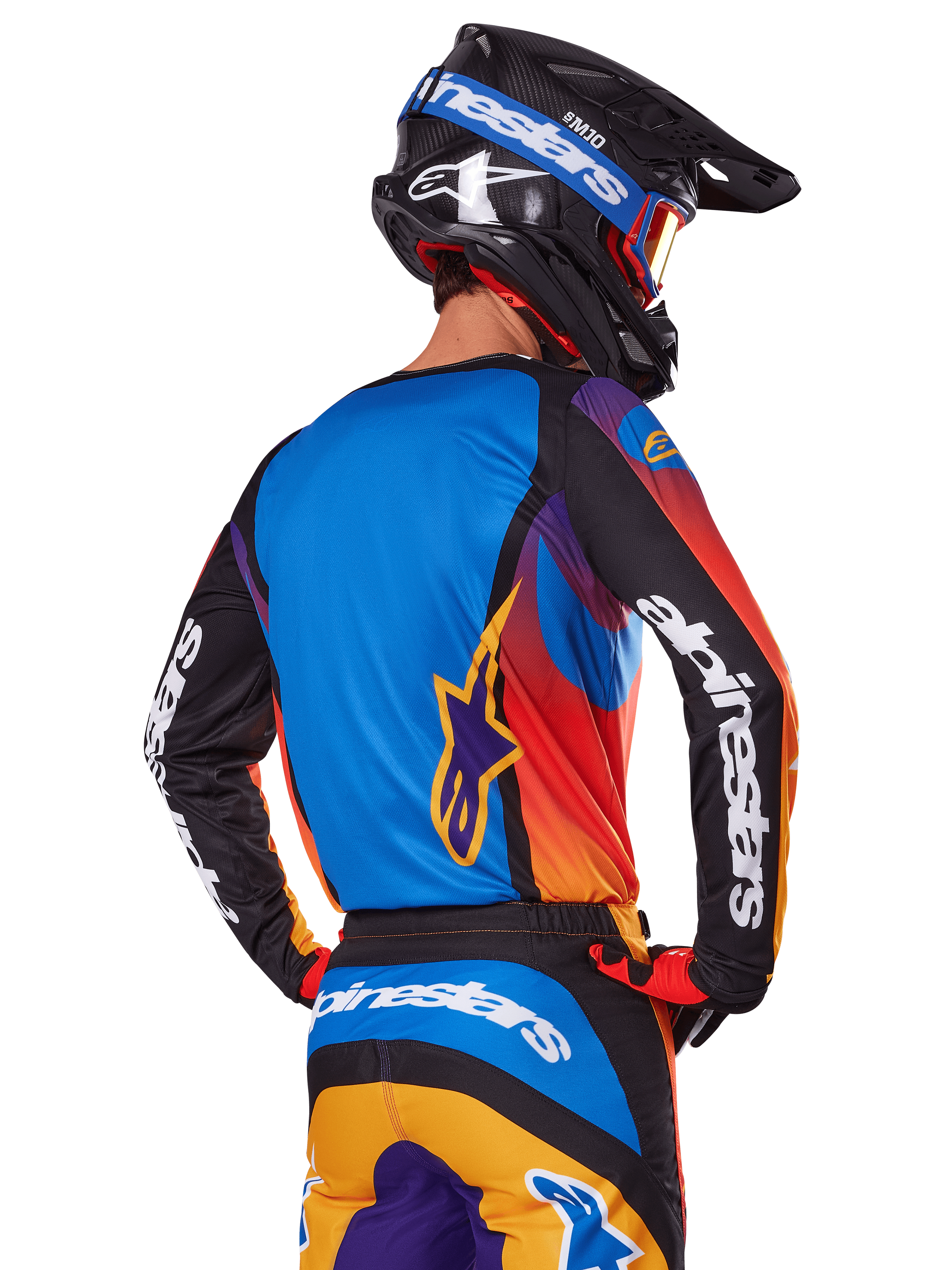 Pilote de motocross vu de dos portant l'ensemble d'équipement Alpinestars 2025 Fluid Wurx, maillot multicolore avec dos bleu et manches noires arborant des logos blancs, pantalon assorti orange et bleu, casque noir et bleu avec lunettes orange, tenue de course tout-terrain coordonnée.