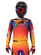 Maillot Alpinestars Fluid Wurx 2025, maillot de motocross multicolore avec dégradé orange et rouge et accents violets, porté avec une tenue assortie, casque noir avec lunettes bleues et rouges et gants rouges, arborant un grand logo étoile noir sur la poitrine.