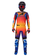 Pilote de motocross portant l'ensemble d'équipement Alpinestars Fluid Wurx 2025, avec un dégradé orange, jaune et bleu vif sur le maillot et le pantalon, logos Alpinestars noirs, assorti à des bottes de course bleues, des gants rouges et noirs, et un casque noir avec lunettes rouges, tenue complète coordonnée pour le tout-terrain