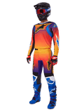 Ensemble d'équipement Alpinestars Fluid Wurx 2025, maillot et pantalon de motocross avec dégradé orange, rouge et violet et motifs étoiles bleues et jaunes, porté avec un casque noir, des lunettes rouges, des gants rouges et noirs et des bottes bleues, tenue de course haute visibilité