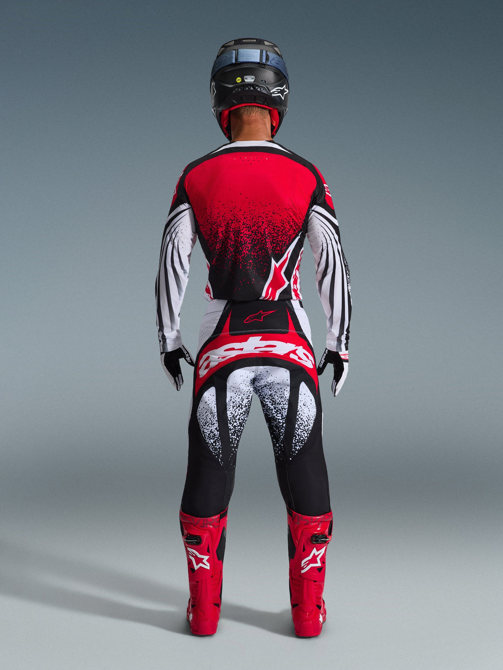 Motocross rider from behind wearing the Alpinestars 2026 Techstar Nomur Jersey, Red Black and White with stippled pattern, matching racing pants, red motocross boots and black helmet, complete gear set for off-road competition