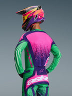 Pilote de motocross vu de dos portant l'ensemble d'équipement Alpinestars 2026 Techstar Nomur, maillot et pantalon assortis violet foncé, vert et rose fluo, avec graphismes audacieux et coupe athlétique, accompagné d'un casque multicolore et de lunettes jaunes.