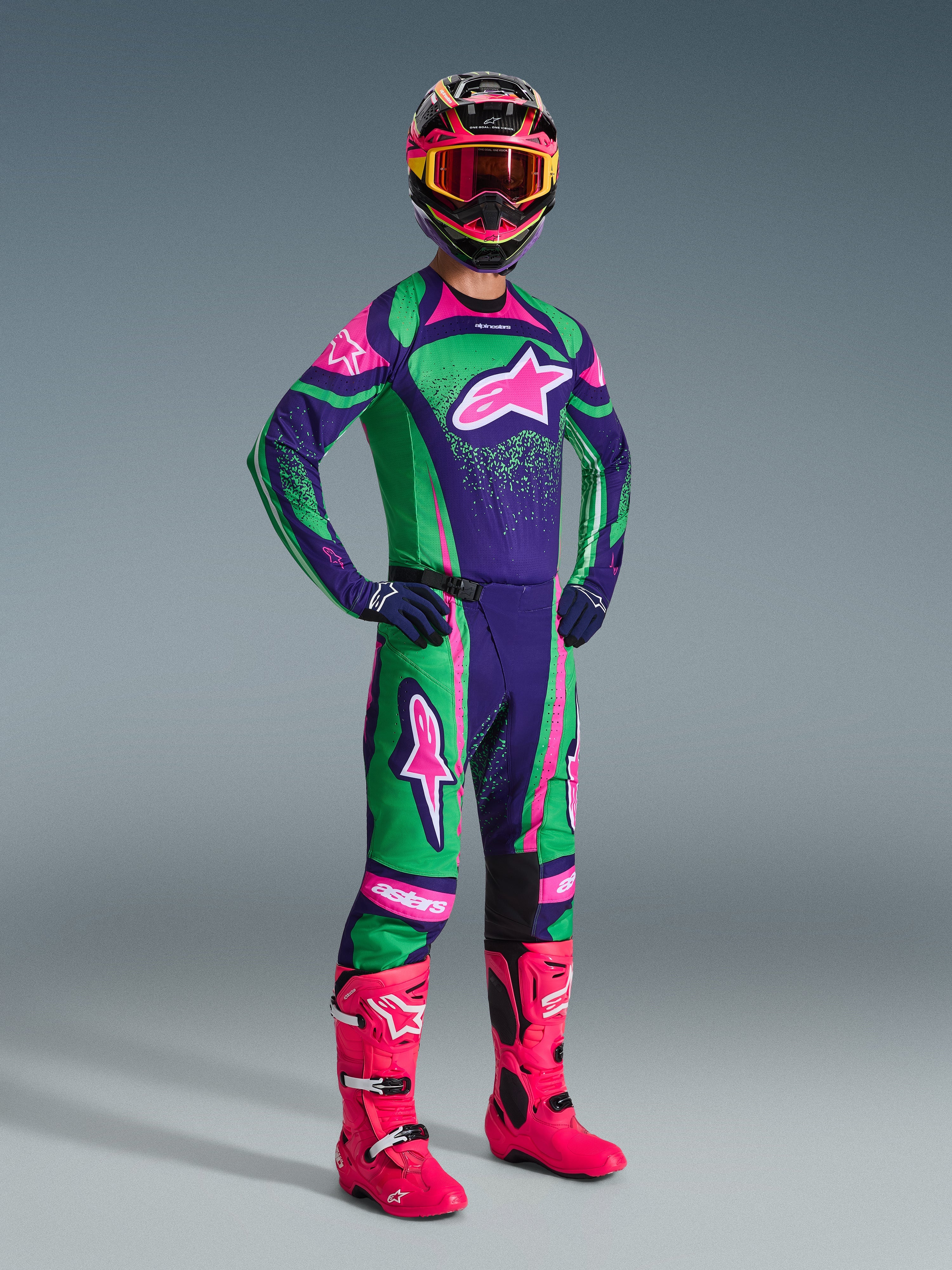 Pilote de motocross portant l'ensemble Alpinestars Techstar Nomur 2026 en violet foncé, vert et rose fluo, comprenant un maillot et un pantalon assortis, un casque coloré avec des lunettes roses et des bottes rose vif, équipement professionnel de course tout-terrain.