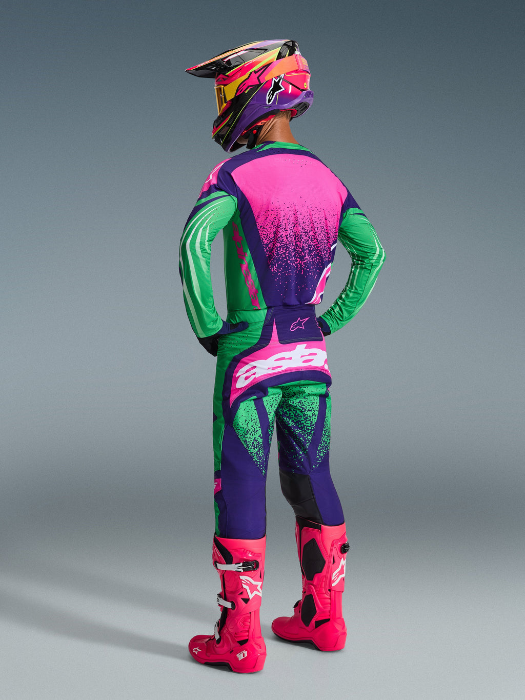 Pilote de motocross portant l'ensemble d'équipement Techstar Nomur 2026, violet foncé, vert et rose fluo, maillot avec dos rose et texture mouchetée, pantalon assorti avec logos blancs, bottes rose fluo vif et casque coloré, vue de dos complète pour la performance en course tout-terrain