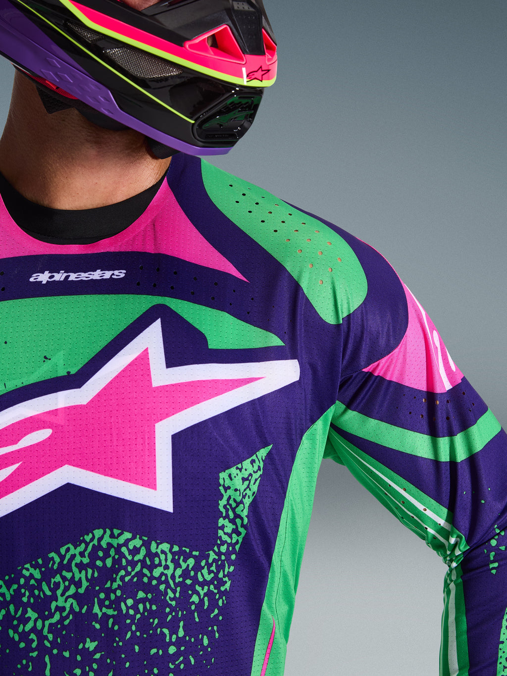 Gros plan sur le maillot Alpinestars Techstar Nomur 2026, violet foncé avec des touches vert et rose fluo, arborant un grand logo étoile rose sur la poitrine et des perforations découpées au laser sur l'épaule, porté par un pilote avec un casque de motocross assorti violet et rose.