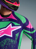 Gros plan sur le maillot Alpinestars Techstar Nomur 2026, violet foncé avec des touches vert et rose fluo, arborant un grand logo étoile rose sur la poitrine et des perforations découpées au laser sur l'épaule, porté par un pilote avec un casque de motocross assorti violet et rose.