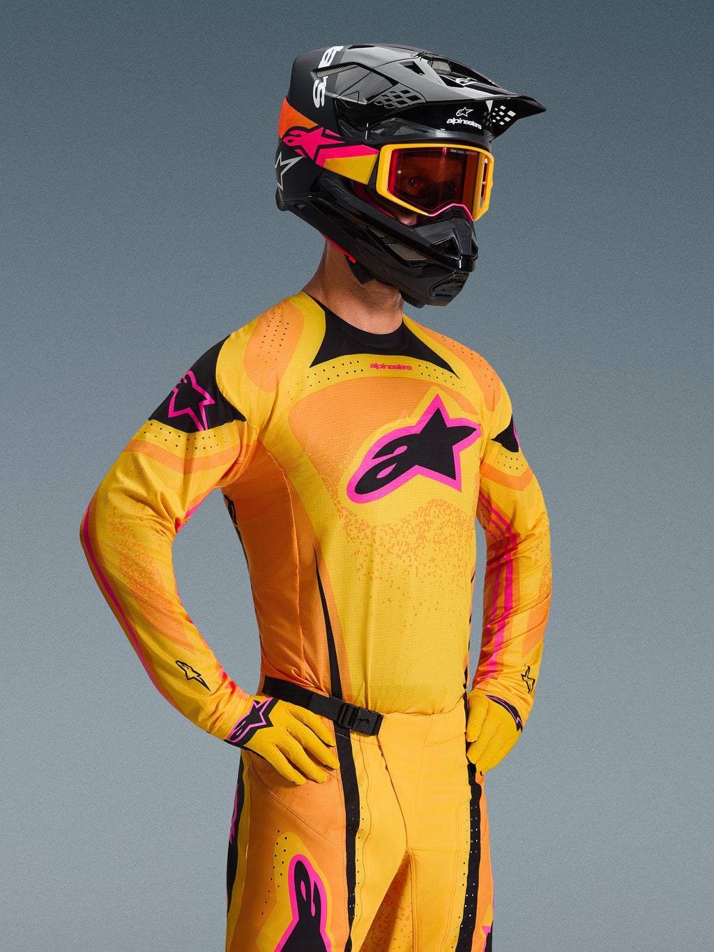 Alpinestars 2026 Techstar Nomur Trikot, Motocross-Trikot, Orange, Gelb und Pink, getragen von einem Fahrer mit passendem schwarzem Helm und Goggles, mit einem großen pinkfarbenen Stern-Logo auf der Brust, schwarzen Akzenten und lasergeschnittenen Belüftungsdetails.