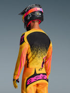 Alpinestars 2026 Techstar Nomur Trikot in Orange-Gelb-Pink, Rückansicht eines Motocross-Fahrers in kompletter Ausrüstung inklusive passender Hose und schwarzem Helm, lebhaftes Design mit gelben und pinkfarbenen Akzenten und schwarzem Rückenpanel, athletische Rennpassform für Offroad-Performance
