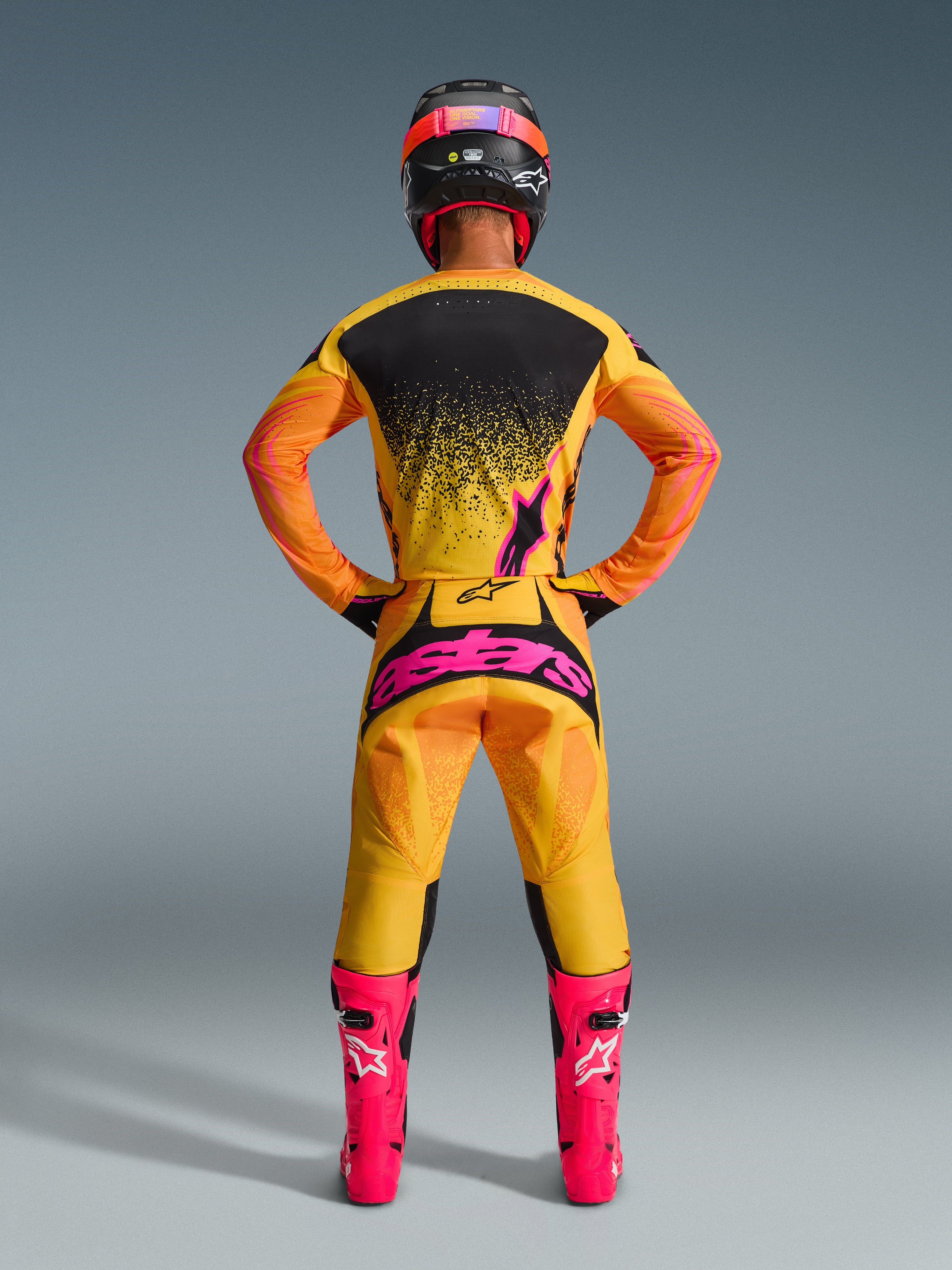 Motocross-Fahrer von hinten im 2026 Techstar Nomur Bekleidungsset in Orange Gelb Pink, bestehend aus einem Trikot mit schwarz-pinken Grafikmustern, passender Hose mit großem pinkem Alpinestars-Branding, leuchtend pinken Rennstiefeln und einem schwarzen Helm mit pinken Akzenten, professionelle Offroad-Rennausrüstung