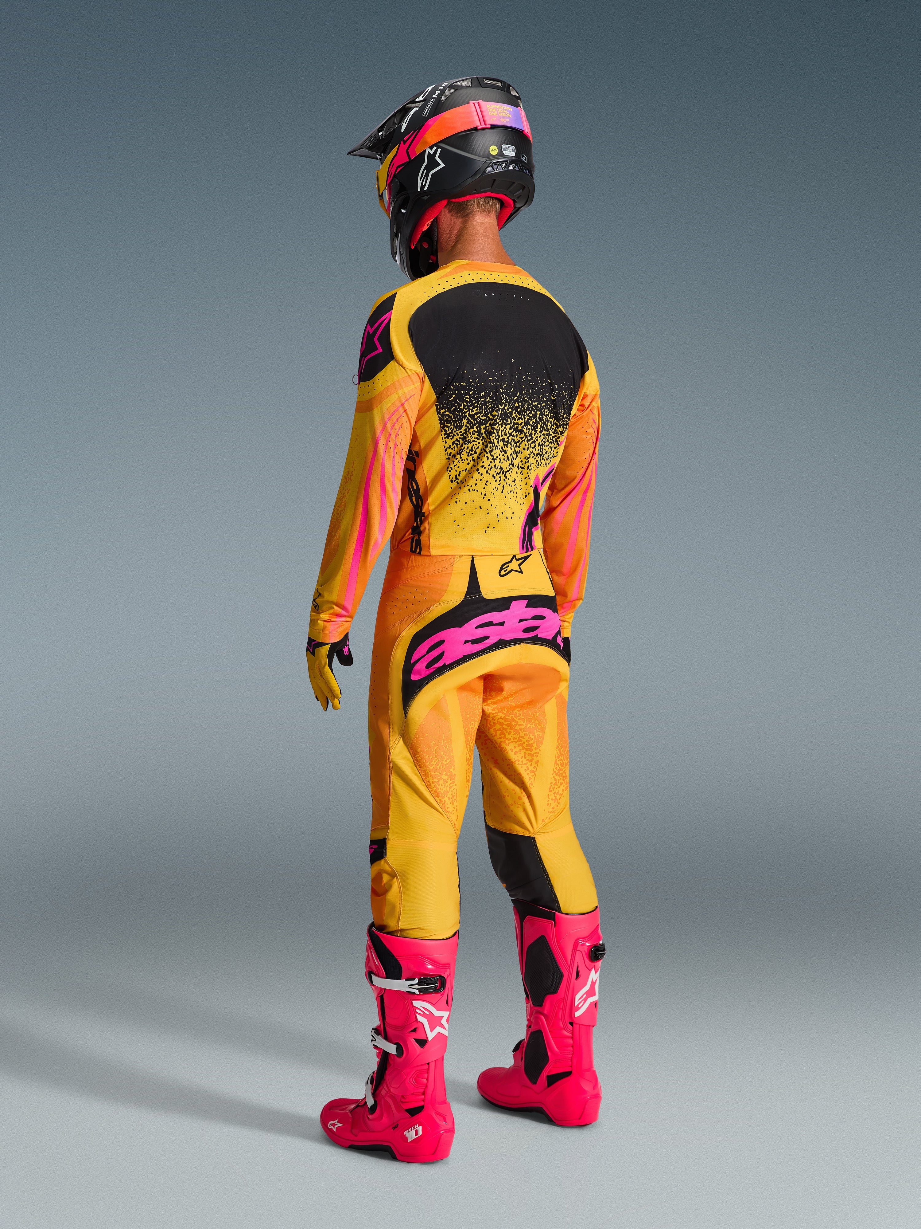 Motocross-Fahrer in der Rückansicht im Alpinestars 2026 Techstar Nomur Gear Set, leuchtendes Trikot in Orange, Gelb und Pink mit schwarzem Grafik-Rückenteil, passende Hose mit auffälligem pinken Alpinestars-Logo auf dem Gesäß, leuchtend pinke Stiefel und schwarzer Rennhelm, koordinierte professionelle Motocross-Rennausrüstung.