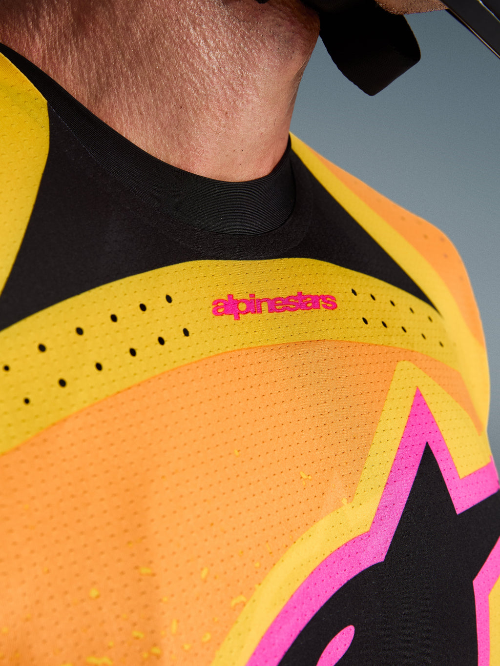 Nahaufnahme des Alpinestars 2026 Techstar Nomur Trikots, Farbkombination Orange, Gelb und Pink, mit lasergeschnittenen Belüftungsperforationen, nahtlosem schwarzem Kragen und pinkfarbenen Branding-Details auf perforiertem Stretchgewebe.