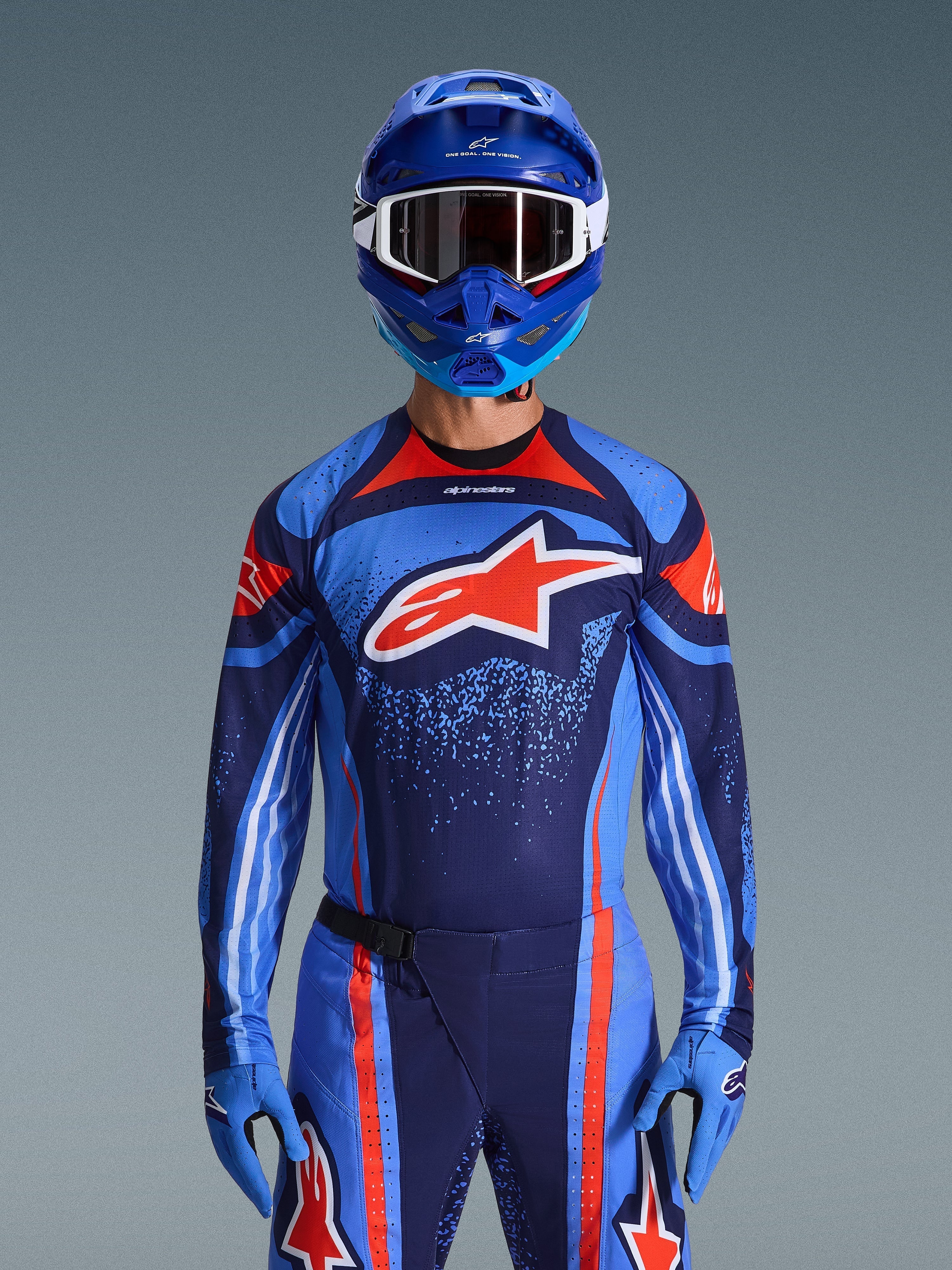 Pilote de motocross portant le maillot Alpinestars Techstar Nomur 2026 en bleu marine foncé, orange et bleu UCLA, avec pantalon assorti et gants bleus, casque bleu avec lunettes blanches, large logo Alpinestars rouge et blanc sur la poitrine, tenue de performance coordonnée pour la course tout-terrain.