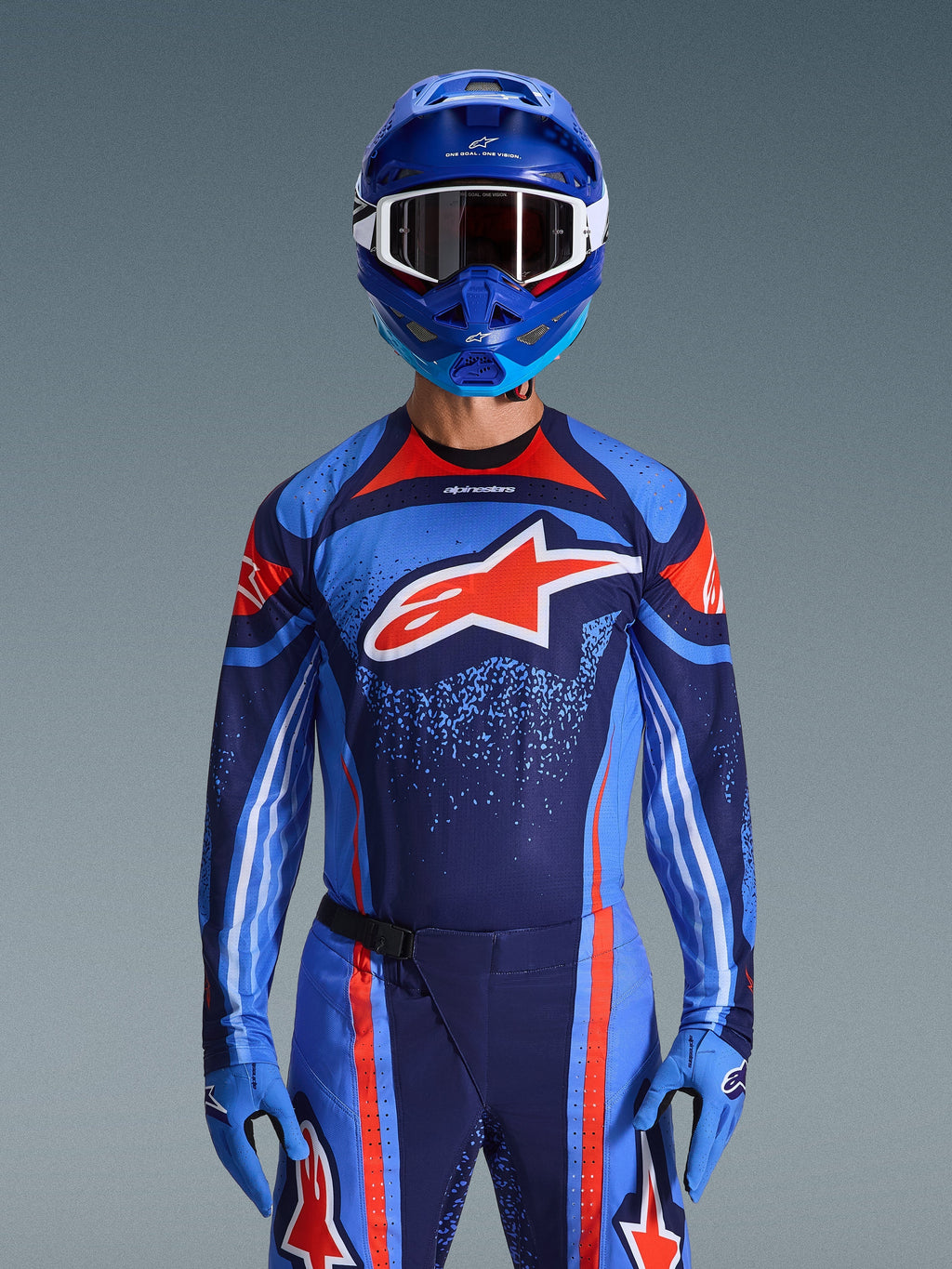 Maillot Alpinestars Techstar Nomur 2026, maillot de motocross, bleu marine foncé, orange et bleu UCLA, porté avec un pantalon assorti, un casque et des gants bleus, arborant un logo étoile blanche sur la poitrine avec des lignes dynamiques et des motifs mouchetés