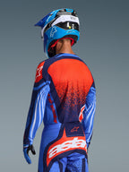 Pilote de motocross vu de dos portant le maillot Alpinestars Techstar Nomur 2026 et le pantalon assorti, bleu marine foncé et bleu UCLA avec des accents orange vifs, présentant un grand graphisme d'étoile et des ventilations découpées au laser sur le maillot, associé à un casque bleu et blanc pour une tenue de course tout-terrain complète.