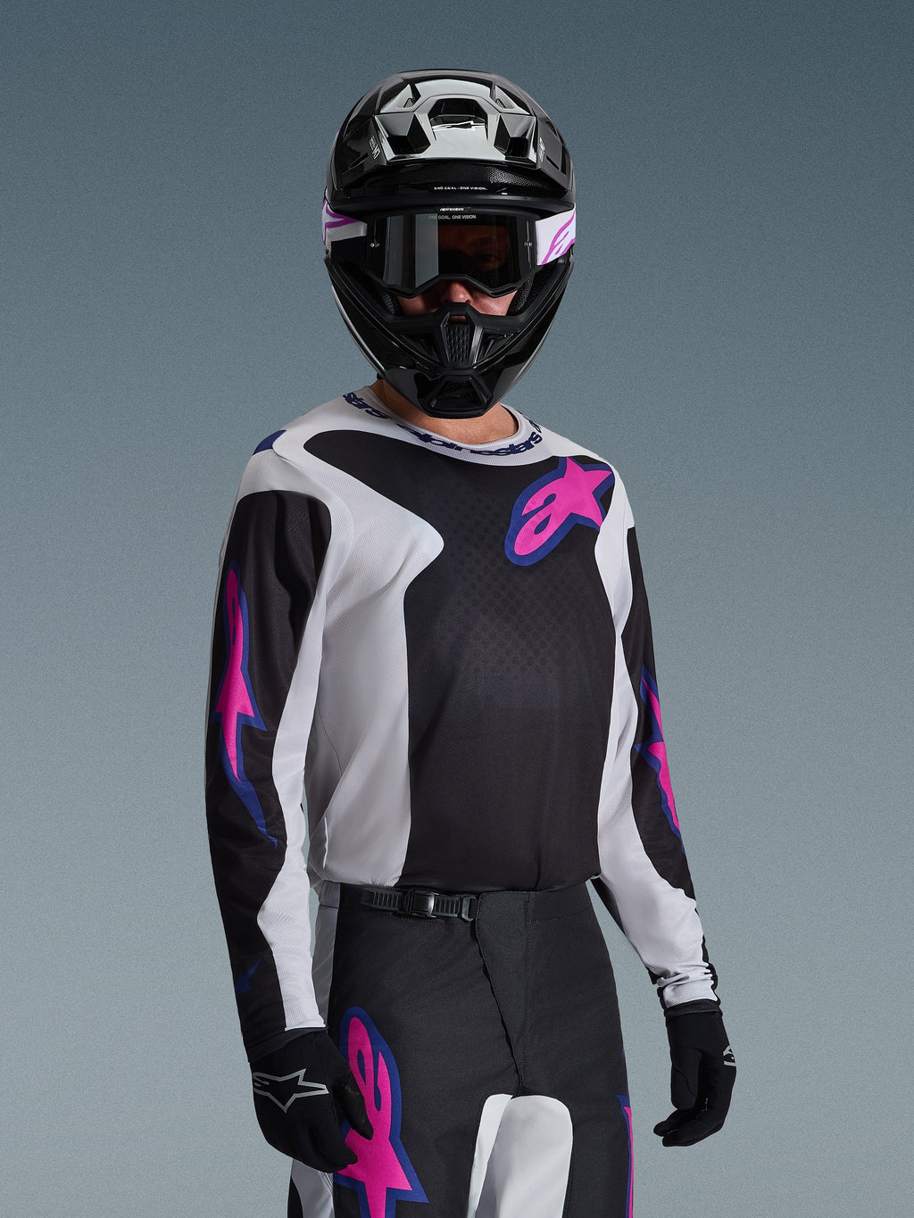 Motocross rider wearing the Alpinestars 2026 Fluid Grid Jersey, Black, Light Gray, and Purple, featuring large pink and purple star logos, paired with matching pants, black helmet, and goggles, complete off-road racing gear set