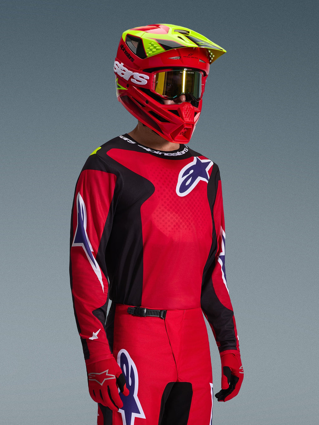 Pilote de motocross portant l'ensemble d'équipement Alpinestars Fluid Grid 2026, maillot rouge et noir avec des accents de logo blancs et bleus, pantalon assorti rouge et noir, casque rouge avec des touches de jaune et des lunettes dorées, équipement de course tout-terrain coordonné
