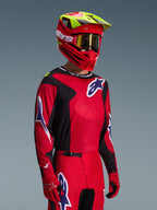 Pilote de motocross portant l'ensemble d'équipement Alpinestars Fluid Grid 2026, maillot rouge et noir avec des accents de logo blancs et bleus, pantalon assorti rouge et noir, casque rouge avec des touches de jaune et des lunettes dorées, équipement de course tout-terrain coordonné