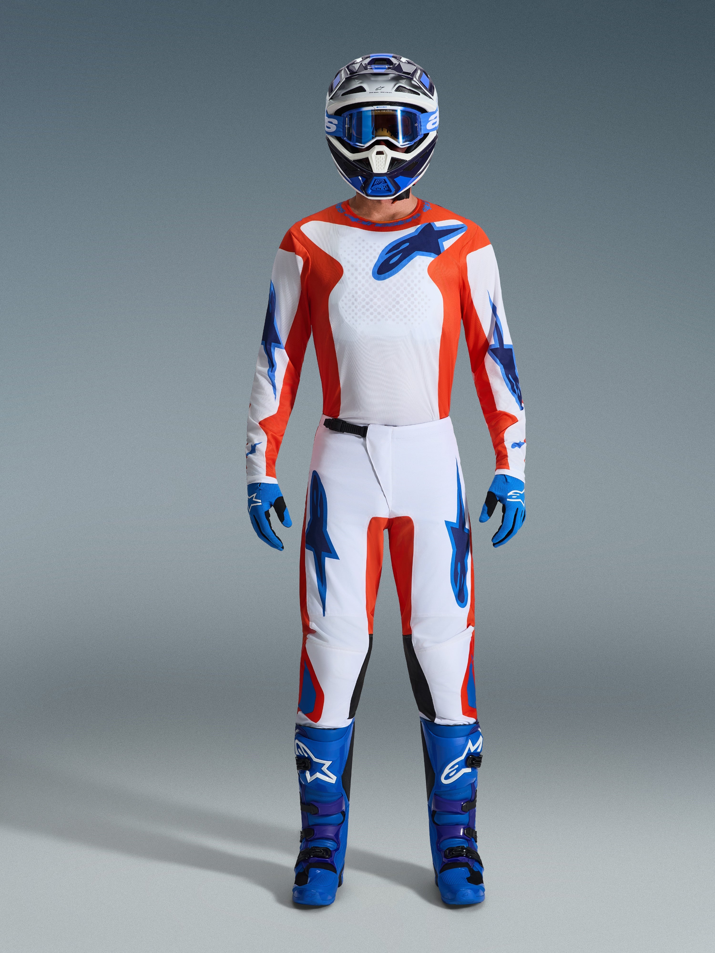 Alpinestars 2026 Fluid Grid Gear Set, pilote de motocross portant un équipement complet, maillot et pantalon blancs avec accents orange et bleu UCLA, casque assorti bleu et blanc avec lunettes bleues, gants bleus et bottes de motocross bleues, tenue de course tout-terrain coordonnée