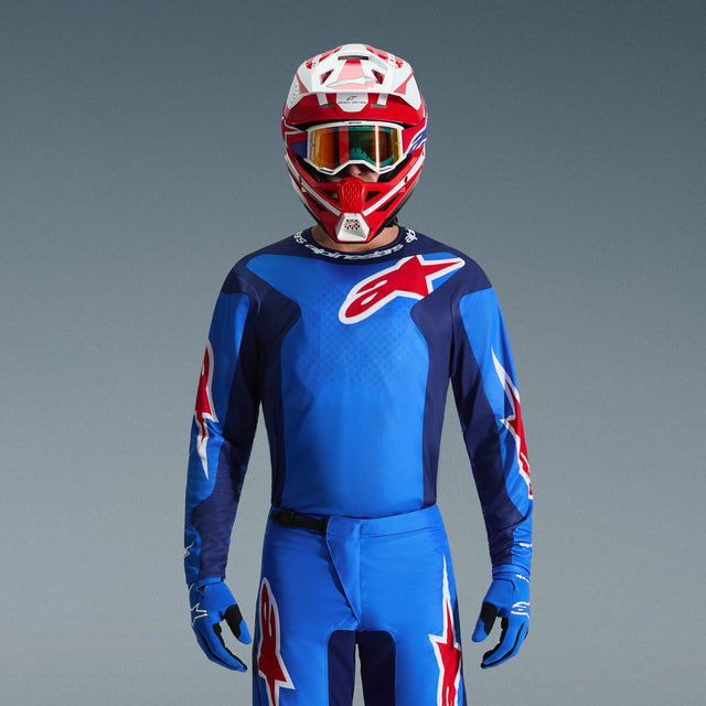 Motocross rider wearing Alpinestars 2026 Fluid Grid Gear Set, bright blue jersey with navy accents and red star logos, matching blue pants, red and white helmet with orange goggles, blue gloves, coordinated outfit for off-road racing